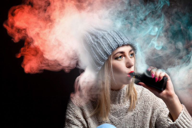 Blonde girl in a knitted sweater and hat holds an electronic cigarette vape in her hand with a manicure and exhales a lot of colored red and green smoke, with red lipstick on a black background.