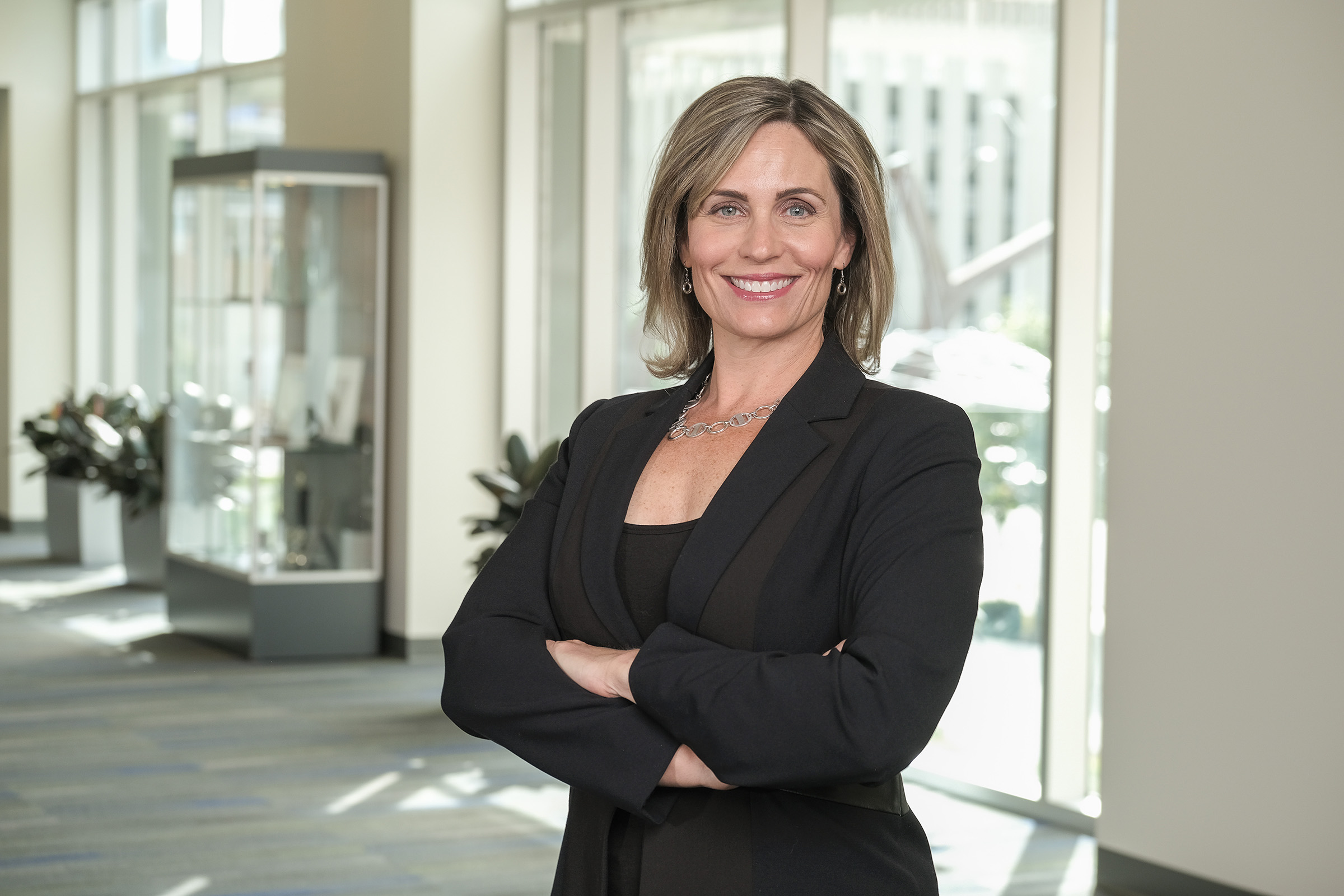 Dr. Nicole Fowler with shoulder-length light brown hair stands with her arms crossed, smiling at the camera. She is wearing a black blazer and a silver necklace. The background features a bright, modern office space with large windows and display cases.